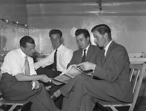 Four men dressed in formal suits are seated in a small, confined room. They appear to be engaged in a serious conversation, with one man holding a newspaper and another pointing at it. The room has a minimalist design, with a few items placed on shelves and tables in the background.