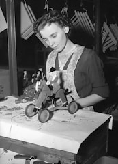 A woman wearing a dark dress with an apron is standing at a workbench assembling a toy horse on wheels. In the background, various items and hanging objects can be seen. The setting appears to be indoors, possibly a workshop or a production area.