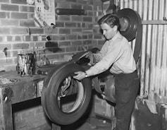 A person is seen working in a workshop environment, handling a large tire. The workspace has brick walls and a wooden workbench covered with various tools and paint cans. There's a roller-like machine or tool mounted on the wall. Various stains and marks are visible on the walls and the tabletop.