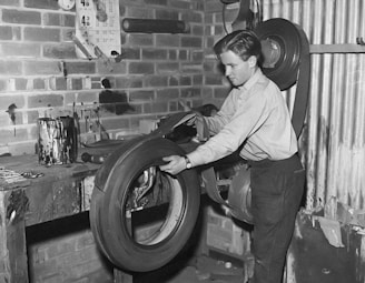 A person is seen working in a workshop environment, handling a large tire. The workspace has brick walls and a wooden workbench covered with various tools and paint cans. There's a roller-like machine or tool mounted on the wall. Various stains and marks are visible on the walls and the tabletop.