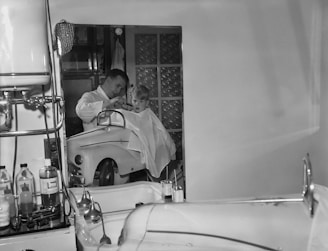 A young child is seated in a chair resembling a toy car while getting a haircut from a barber. The barber appears focused, wearing a white coat, and the room includes various equipment typically found in a barbershop. The decor suggests a mid-20th-century setting, with a mix of chrome and glass elements.