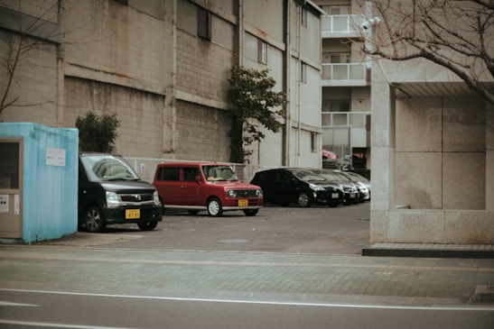 A parking lot adjacent to a tall building with multiple cars parked. The cars include a black van, a red compact car, and several other black sedans. There is an old blue booth structure to the left of the image and a tree without leaves on the right. The surrounding area looks like an urban environment with concrete walls and buildings.