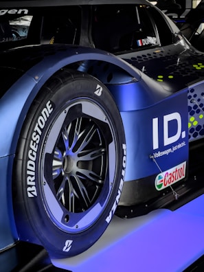 A close-up of a car's rear wheel featuring Bridgestone Potenza tires. The car has a sleek, modern design with a blue and black color scheme. The logo and branding for Volkswagen ID and Castrol are visible, suggesting it is a high-performance electric vehicle.