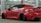 A sleek, customized red sports car is parked indoors. The vehicle features a wide body kit, lowered suspension, and dark tinted windows. It is positioned at an angle, revealing its aggressive styling and prominent rear wheels. The setting appears to be a garage or workshop with industrial lighting and a red beam in the background.