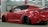 A sleek, customized red sports car is parked indoors. The vehicle features a wide body kit, lowered suspension, and dark tinted windows. It is positioned at an angle, revealing its aggressive styling and prominent rear wheels. The setting appears to be a garage or workshop with industrial lighting and a red beam in the background.