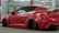 A sleek, customized red sports car is parked indoors. The vehicle features a wide body kit, lowered suspension, and dark tinted windows. It is positioned at an angle, revealing its aggressive styling and prominent rear wheels. The setting appears to be a garage or workshop with industrial lighting and a red beam in the background.