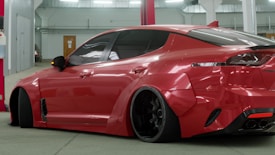 A sleek, customized red sports car is parked indoors. The vehicle features a wide body kit, lowered suspension, and dark tinted windows. It is positioned at an angle, revealing its aggressive styling and prominent rear wheels. The setting appears to be a garage or workshop with industrial lighting and a red beam in the background.