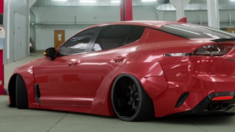 A sleek, customized red sports car is parked indoors. The vehicle features a wide body kit, lowered suspension, and dark tinted windows. It is positioned at an angle, revealing its aggressive styling and prominent rear wheels. The setting appears to be a garage or workshop with industrial lighting and a red beam in the background.