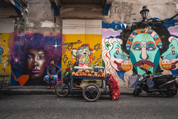 A vibrant street scene features colorful murals on walls, including a striking portrait of a woman with an afro and bold makeup, alongside imaginative abstract designs. A street vendor sits in front of the murals, beside a cart filled with coconuts and a sack of onions. A motorbike is parked nearby.