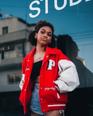 A young woman stands confidently outdoors wearing a red varsity jacket with a prominent 'P' patch on the left side, a black crop top, and denim shorts. The background includes a reflective glass window with part of the word 'STUD...' visible, and an urban structure with windows.