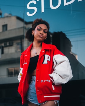 A young woman stands confidently outdoors wearing a red varsity jacket with a prominent 'P' patch on the left side, a black crop top, and denim shorts. The background includes a reflective glass window with part of the word 'STUD...' visible, and an urban structure with windows.