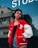 A young woman stands confidently outdoors wearing a red varsity jacket with a prominent 'P' patch on the left side, a black crop top, and denim shorts. The background includes a reflective glass window with part of the word 'STUD...' visible, and an urban structure with windows.