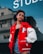 A young woman stands confidently outdoors wearing a red varsity jacket with a prominent 'P' patch on the left side, a black crop top, and denim shorts. The background includes a reflective glass window with part of the word 'STUD...' visible, and an urban structure with windows.