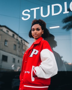 A person with curly hair wearing a red and white varsity jacket with the letter P on the chest, standing in front of a glass window with a reflection and the word 'STUDIO' in large letters above. The background shows the facade of an older building and a palm tree reflected in the glass.