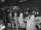 A group of young men and one woman are seated in a sewing workshop, with sewing machines and spools of thread visible. The men are wearing work attire and are seated in front of machines, seemingly involved in textile work. The setting appears to be a vocational or educational environment.
