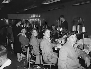 A group of young men and one woman are seated in a sewing workshop, with sewing machines and spools of thread visible. The men are wearing work attire and are seated in front of machines, seemingly involved in textile work. The setting appears to be a vocational or educational environment.