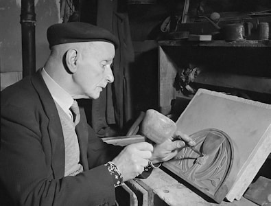 An elderly man wearing a beret and a suit is engaged in intricate carving or engraving work on a rectangular piece of material. He holds a tool in one hand and a mallet in the other. The room appears to be a workshop, with various tools and objects scattered in the background.