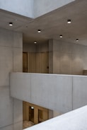 A modern architectural space with concrete walls and ceilings. The design is minimalist with clean lines and subtle lighting fixtures affixed to the ceiling. There are metal elements visible, such as doors or panels, likely made from brushed brass or similar material, contributing to the contemporary aesthetic.