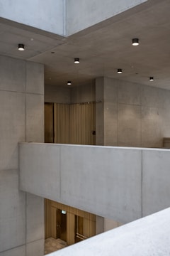 A modern architectural space with concrete walls and ceilings. The design is minimalist with clean lines and subtle lighting fixtures affixed to the ceiling. There are metal elements visible, such as doors or panels, likely made from brushed brass or similar material, contributing to the contemporary aesthetic.