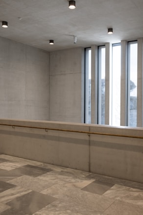 A minimalist interior space with concrete walls and ceiling, featuring several recessed ceiling lights. Large vertical windows allow natural light to enter, casting shadows on the polished stone floor. A simple railing is attached to the wall.