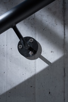 A sleek black handrail is mounted on a textured concrete wall. The handrail is attached with a metal fixture, which casts a sharp shadow across the wall. The lighting creates contrasts between the dark handrail and the lighter wall surface.