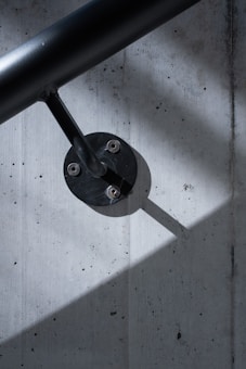 A sleek black handrail is mounted on a textured concrete wall. The handrail is attached with a metal fixture, which casts a sharp shadow across the wall. The lighting creates contrasts between the dark handrail and the lighter wall surface.