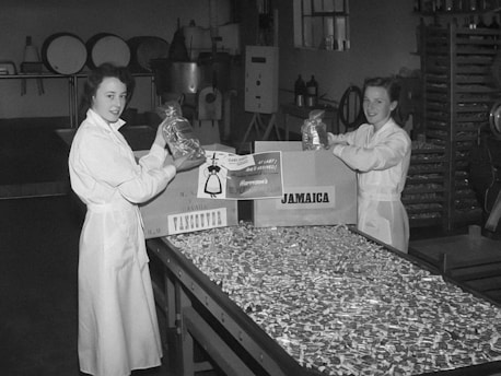 Two women wearing white lab coats are standing in a factory setting, holding bags of candies or goods. A large table covered with numerous small packages is in front of them. Boxes labeled 'Vancouver' and 'Jamaica' are nearby, suggesting packaging or distribution activities. The background features industrial equipment and shelves.