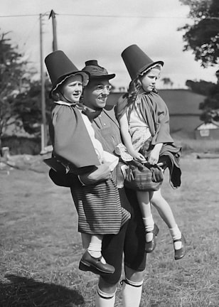 A person dressed in traditional Dutch clothing holds two young children, also in similar attire. The background features a rural setting with trees and a building.