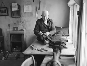 An elderly man wearing a suit is sitting cross-legged on a large wooden table while working on fabric with a needle and thread. The room appears somewhat aged, featuring a calendar and a clock on the wall, as well as several tools scattered on the table. The atmosphere is serene and focused.