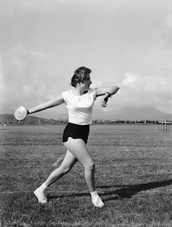 A woman is in mid-action preparing to throw a discus in an open grassy field. She is wearing a short-sleeved shirt, shorts, and athletic shoes. The background features a distant view of mountains and a vast sky with scattered clouds.