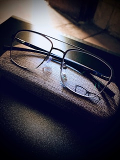A pair of eyeglasses rests on a textured glasses case on a dark, reflective surface. The lighting creates a contrast, highlighting the clarity of the lenses against the muted tones of the background.