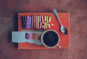 An artistic arrangement featuring a collection of pastel chalks in shades of pink, purple, orange, and yellow neatly lined up on a textured surface. A small piece of paper displays sample color strokes matching the chalks. A vintage silver spoon and a cup of dark liquid, possibly coffee or tea, accompany this creative setup. The background is a warm, earthy brown surface.