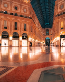 An elegant, illuminated indoor shopping gallery with a glass-vaulted ceiling and ornate architectural details. Luxury brand storefronts like Prada and Louis Vuitton are visible, with a few people walking on the polished marble floor. The lighting creates a warm ambiance.