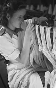 A woman is engaged in a textile-related task, possibly sewing or fabric inspection. She is wearing a white garment and is surrounded by strips of fabric. The setting appears to be a workshop or factory environment.