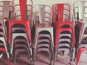 A stack of metal chairs arranged in an orderly manner, with alternating colors of red and metallic gray. The chairs are placed against a neutral-colored wall, and their industrial design is evident.