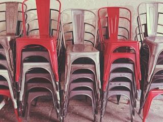 A stack of metal chairs arranged in an orderly manner, with alternating colors of red and metallic gray. The chairs are placed against a neutral-colored wall, and their industrial design is evident.