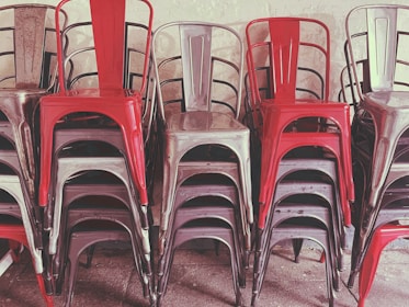 A stack of metal chairs arranged in an orderly manner, with alternating colors of red and metallic gray. The chairs are placed against a neutral-colored wall, and their industrial design is evident.