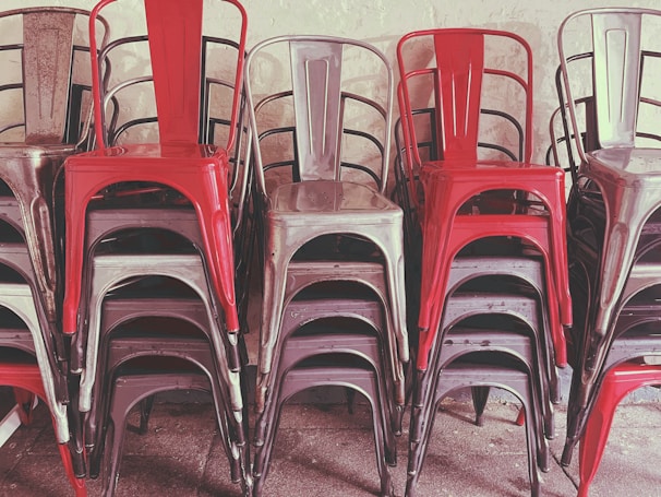 A stack of metal chairs arranged in an orderly manner, with alternating colors of red and metallic gray. The chairs are placed against a neutral-colored wall, and their industrial design is evident.