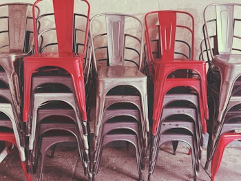 A stack of metal chairs arranged in an orderly manner, with alternating colors of red and metallic gray. The chairs are placed against a neutral-colored wall, and their industrial design is evident.