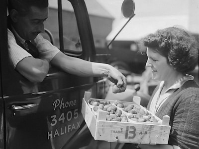 A man in a truck interacts with a woman who is carrying a crate filled with containers of strawberries. The truck has a sign with a phone number and the word 'Halifax' on the door. The woman, smiling, looks at the man as he inspects the produce.