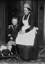 A vintage black and white photograph featuring a woman seated next to a young boy. The woman is dressed in a traditional maid's outfit with an apron and a cap. The boy is wearing a sailor-style suit and is holding a toy horse on wheels. The background consists of a dark wooden door, conveying a sense of formality and historic charm.