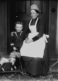 A vintage black and white photograph featuring a woman seated next to a young boy. The woman is dressed in a traditional maid's outfit with an apron and a cap. The boy is wearing a sailor-style suit and is holding a toy horse on wheels. The background consists of a dark wooden door, conveying a sense of formality and historic charm.