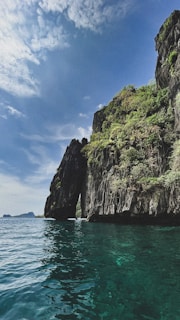 A towering rock formation covered with lush greenery rises dramatically above a turquoise sea. The sky is clear with a few clouds, adding a serene backdrop to the scene.