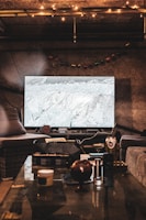 A cozy room with dim lighting, featuring a large television displaying a snowy mountain scene. The room has comfortable seating, including a wicker chair and a sofa. A glass coffee table in the foreground is cluttered with various items such as a can, a jar, and other miscellaneous objects. Warm, string lights hang from the ceiling, providing an inviting ambiance.
