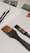 A collection of art supplies laid out on a white surface, including a blank canvas, various paintbrushes with different sizes and bristle colors, and tubes of paint. The brushes vary in size, one with fine tips and another with a wide, orange bristle. Paint tubes are slightly squeezed, indicating use.