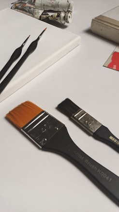 A collection of art supplies laid out on a white surface, including a blank canvas, various paintbrushes with different sizes and bristle colors, and tubes of paint. The brushes vary in size, one with fine tips and another with a wide, orange bristle. Paint tubes are slightly squeezed, indicating use.
