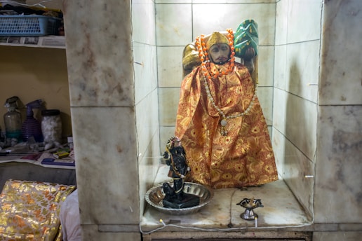 A statue adorned with an ornate golden and red robe sits in a small alcove with white marble tiles. The statue wears a string of orange beads and a decorative headpiece. Nearby, a small black figure is placed inside a silver dish, and a metal oil lamp is seen in front. The surrounding area contains a shelf with miscellaneous objects and cleaning supplies.
