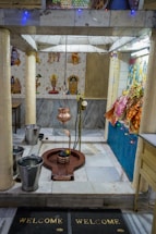 A small temple interior with religious artifacts and decorations. The central object appears to be a Shiva Lingam with a hanging copper vessel above it, likely used for water or milk offerings. The surrounding area is adorned with small statues and colorful cloths. The walls are decorated with tiles featuring images of deities, and there are two buckets placed on the floor.