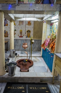A small temple interior with religious artifacts and decorations. The central object appears to be a Shiva Lingam with a hanging copper vessel above it, likely used for water or milk offerings. The surrounding area is adorned with small statues and colorful cloths. The walls are decorated with tiles featuring images of deities, and there are two buckets placed on the floor.