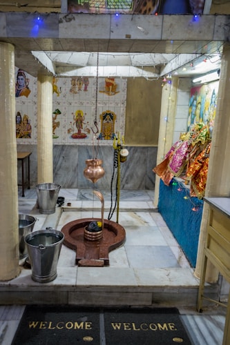 A small temple interior with religious artifacts and decorations. The central object appears to be a Shiva Lingam with a hanging copper vessel above it, likely used for water or milk offerings. The surrounding area is adorned with small statues and colorful cloths. The walls are decorated with tiles featuring images of deities, and there are two buckets placed on the floor.